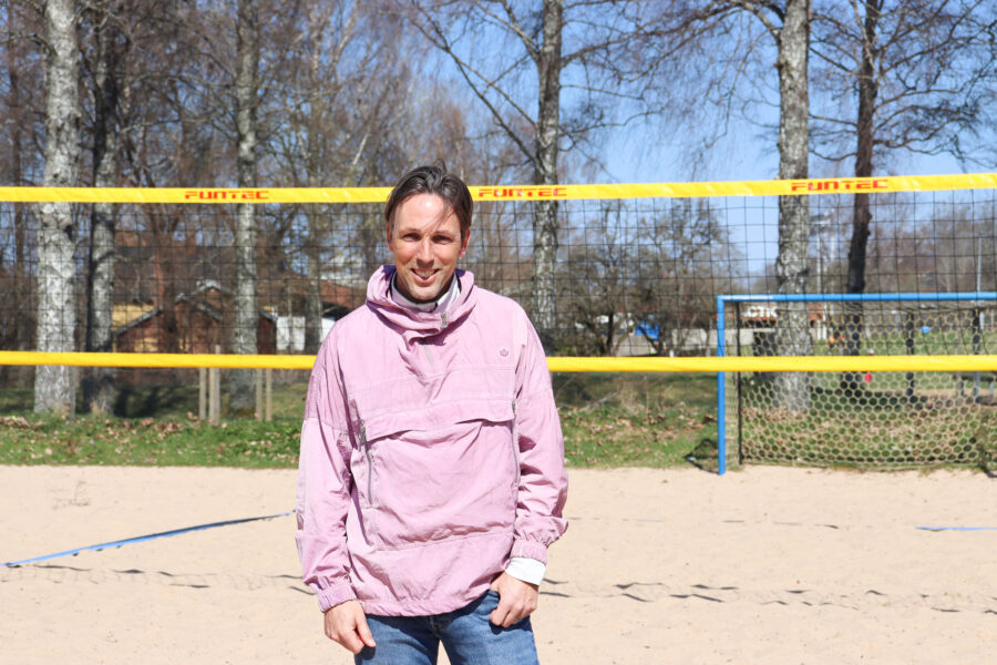 Joakim står i sanden framför ett volleybollnät. Han har rosa jacka och vinden rufsar om hans hår.